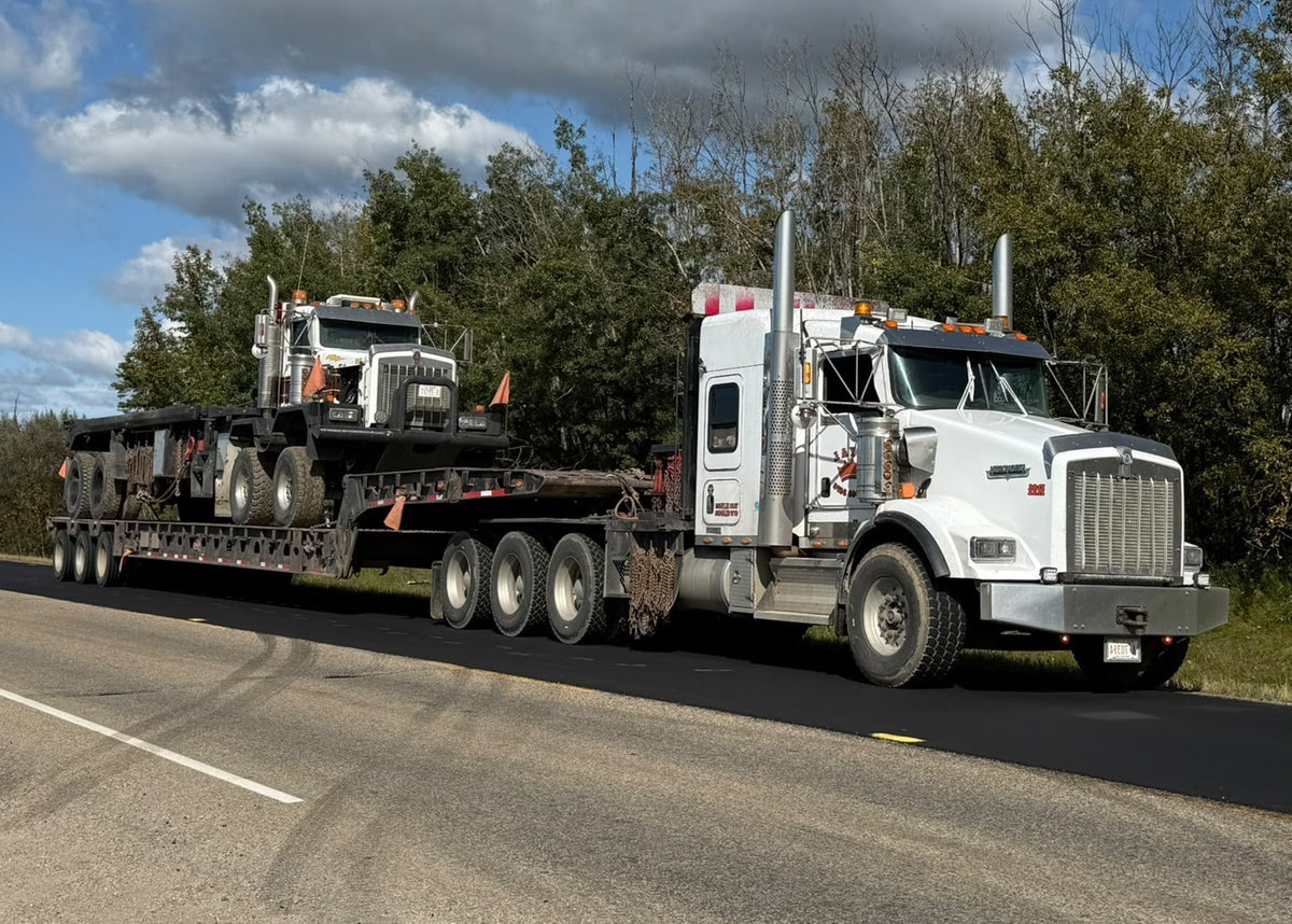 A white semi-truck towing a long flatbed trailer carrying another truck, traveling on a paved road under a blue sky with large white clouds and trees in the background.